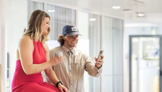 Nainen punaisessa mekossa ja mies lippiksessä katsovat yhdessä puhelinta toimistossa.