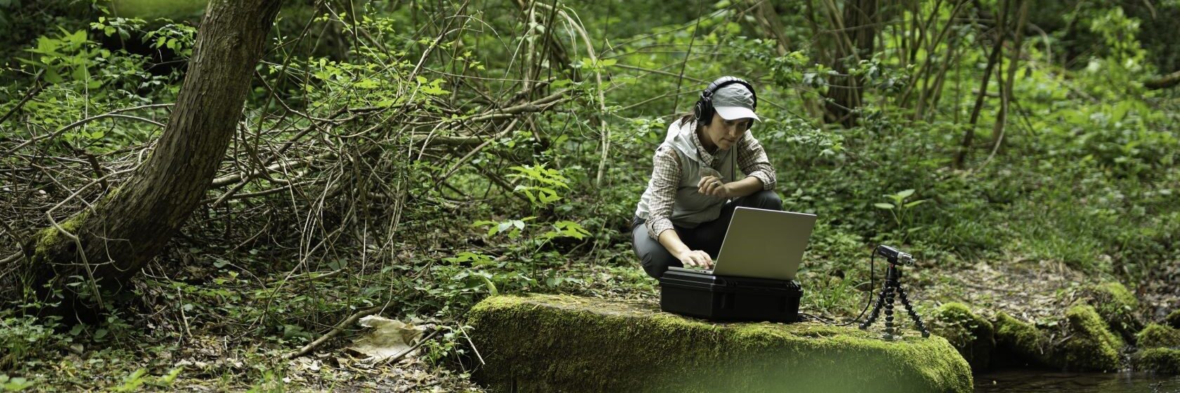 Nainen on kyykyssä kannettavan tietokoneen ja mikrofonin kanssa vehreässä lehtometsässä puron rannalla.