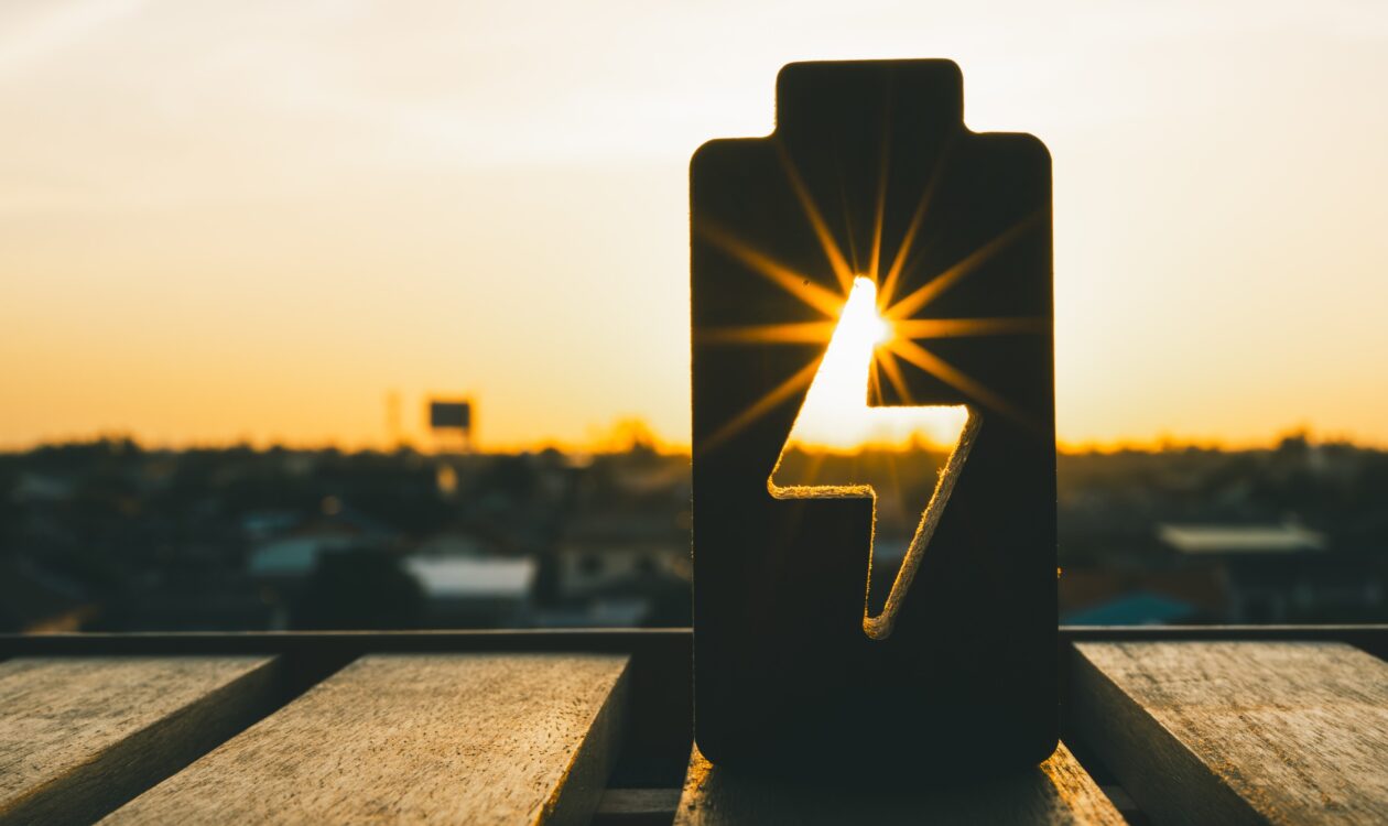 Aurinkoinen maisema, jossa siluettina näkyy akun muotoinen kuvio ja sen keskellä salaman symboli. Aurinko paistaa salaman läpi, luoden kirkkaan valon säteet akun sisään.