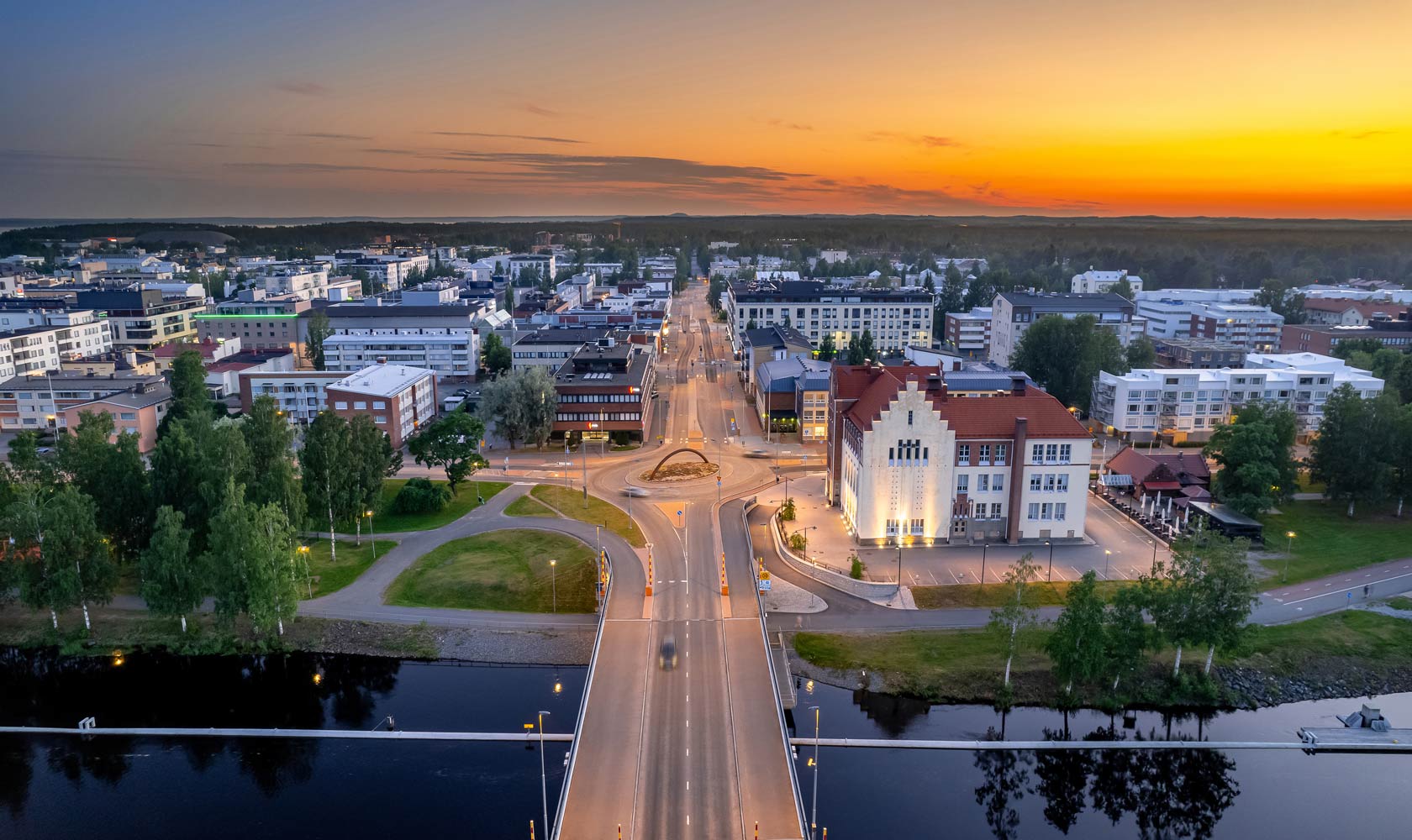 Aerial view of Joensuu city center at sunset, showing a bridge crossing the Pielisjoki River, illuminated streets, historic and modern buildings, and a vibrant urban landscape with trees and waterways