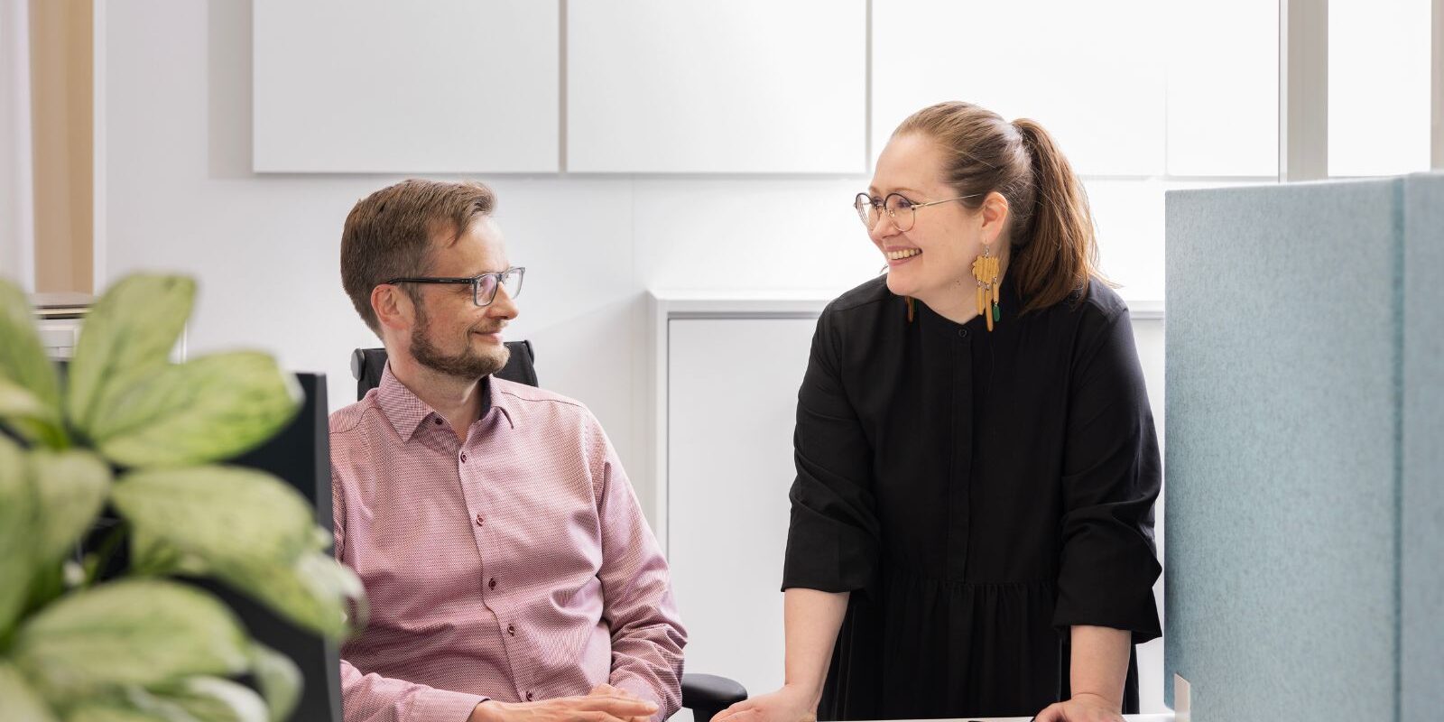 Two colleagues discussing work at a computer desk in a modern office setting.