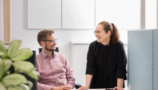 Two colleagues discussing work at a computer desk in a modern office setting.