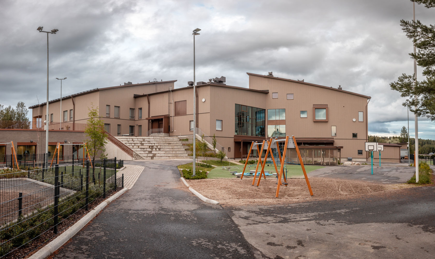 Modern school building with playground swings, basketball court, and cloudy sky background.