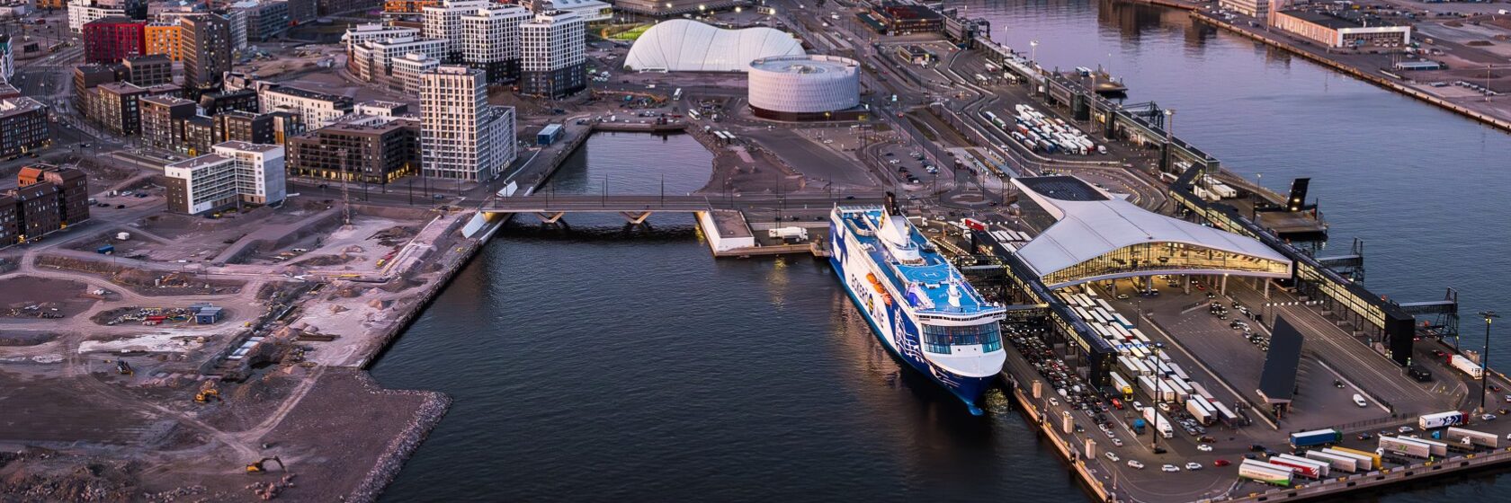 Ilmakuva Helsingin satama-alueesta, laiva, rakennuksia auringonlaskun aikaan.