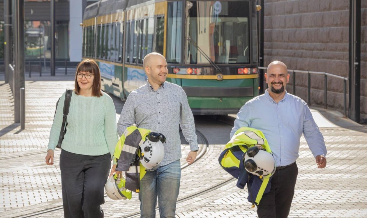 Keski-ikäinen nainen, nuori mies ja keski-ikäinen mies kävelevät neonkeltaiset huomioliivit ja työmaakypärät kainaloissaan, taustalla raitiovaunu