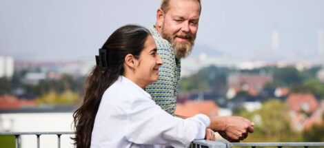 Nainen ja mies keskustelevat ulkona, nojaavat kaiteeseen, taustalla näkyy kaupungin silhuetti