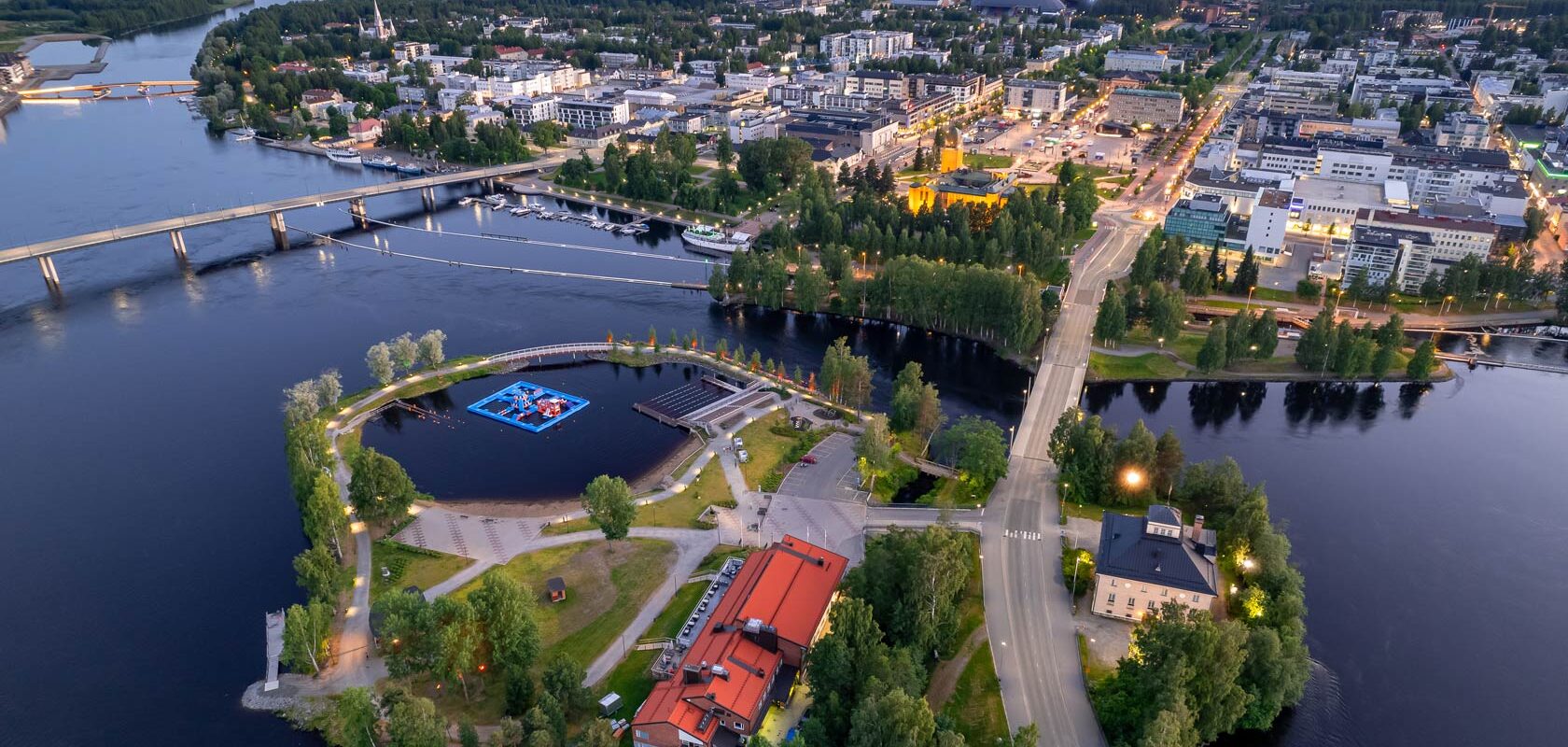 Aerial view of Joensuu, Finland at dusk, showing the city center, bridges crossing the Pielisjoki River, waterfront areas, and green spaces, with buildings and streets illuminated by evening lights.