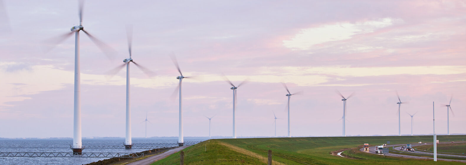 Row of wind turbines along a coastal landscape with a road and vehicles, generating renewable energy under a cloudy sky
