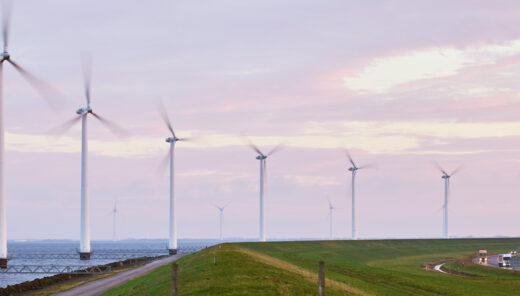 Row of wind turbines along a coastal landscape with a road and vehicles, generating renewable energy under a cloudy sky