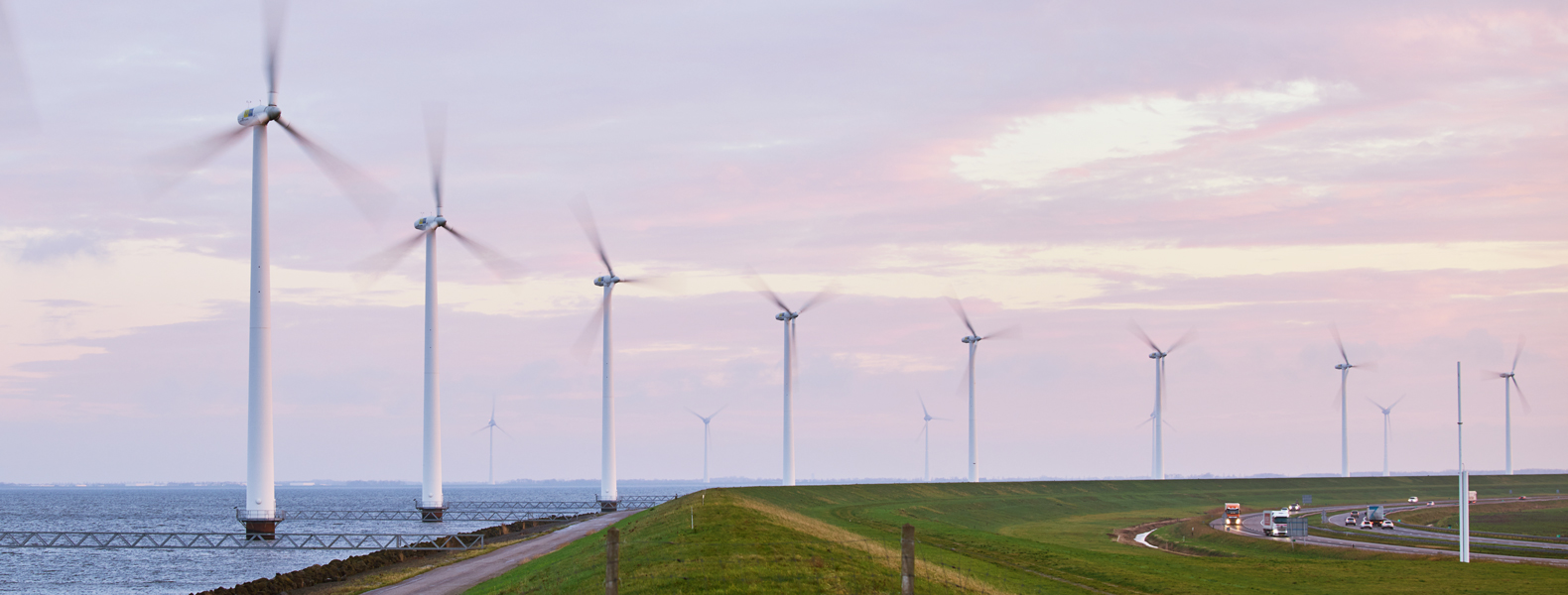 Row of wind turbines along a coastal landscape with a road and vehicles, generating renewable energy under a cloudy sky