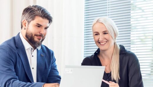 A man and a woman are sitting and smiling in front of a laptop in a bright meeting room.