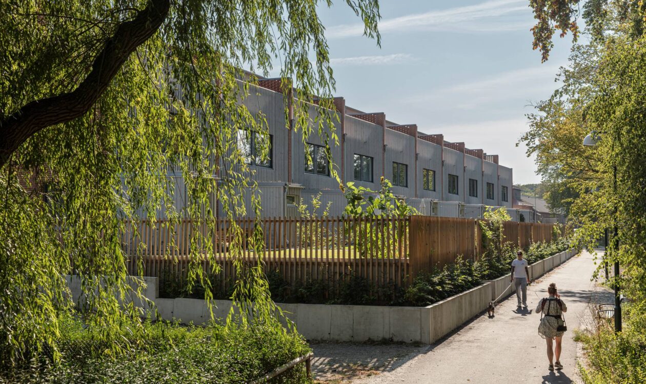 A sunny pedestrian path runs alongside a row of modern, gray townhouses, bordered by wooden fencing and lush greenery, with two people walking along the tree‑lined walkway