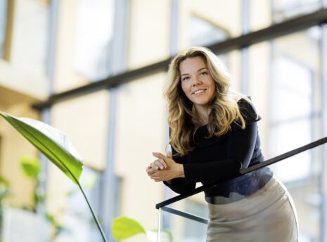 Pitkähiuksinen pohjoiseurooppalainen nainen nojaa lasikaiteeseen valoisassa toimistotilassa.