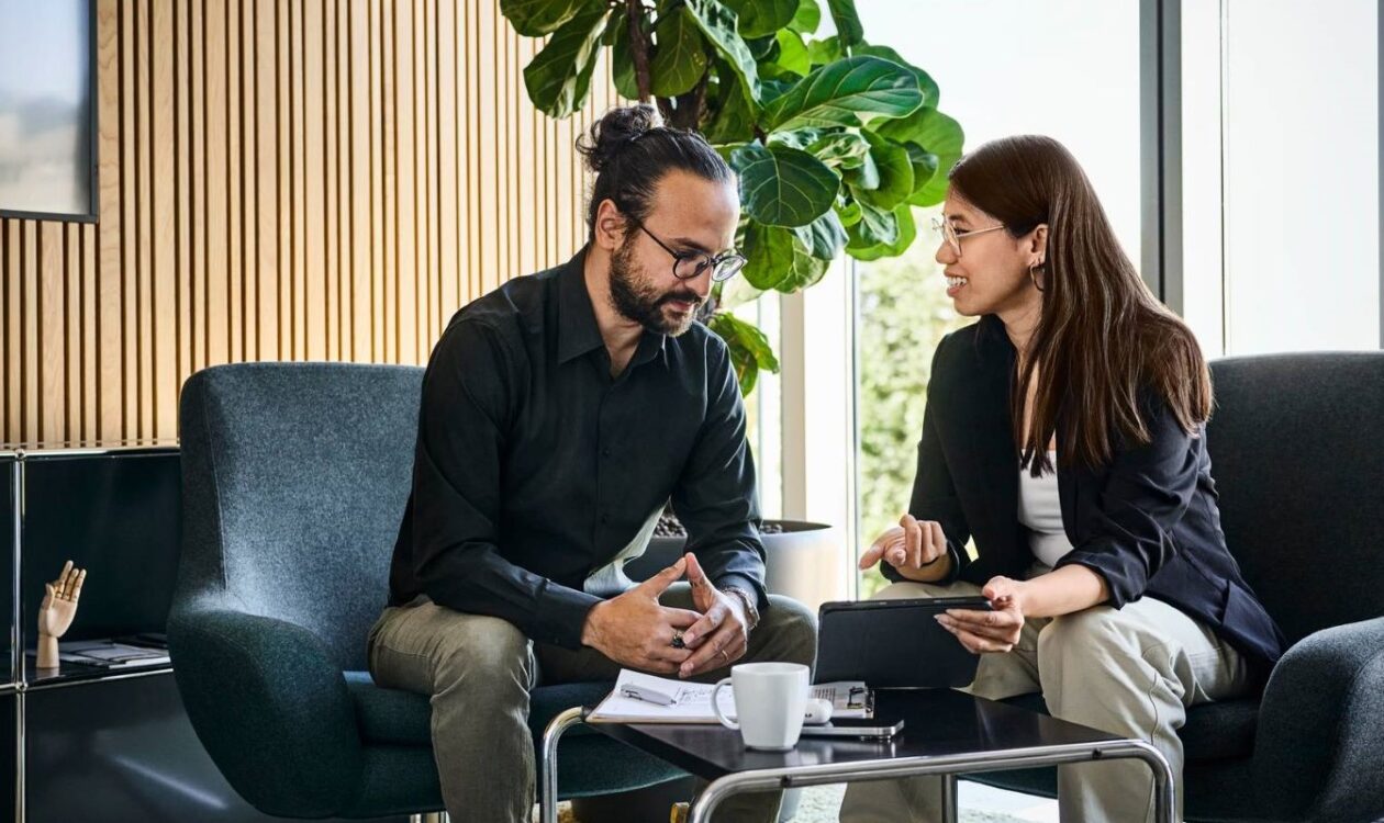 Mies ja nainen keskustelemassa nojatuoleissa modernissa toimistossa, nainen esittelee miehelle padilta asiaansa, miehellä on kynä ja lehtiö edessään matalalla pöydällä.
