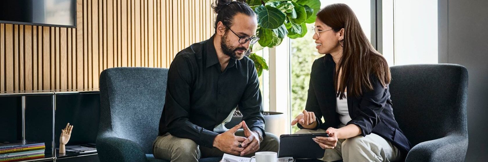 Mies ja nainen keskustelemassa nojatuoleissa modernissa toimistossa, nainen esittelee miehelle padilta asiaansa, miehellä on kynä ja lehtiö edessään matalalla pöydällä.
