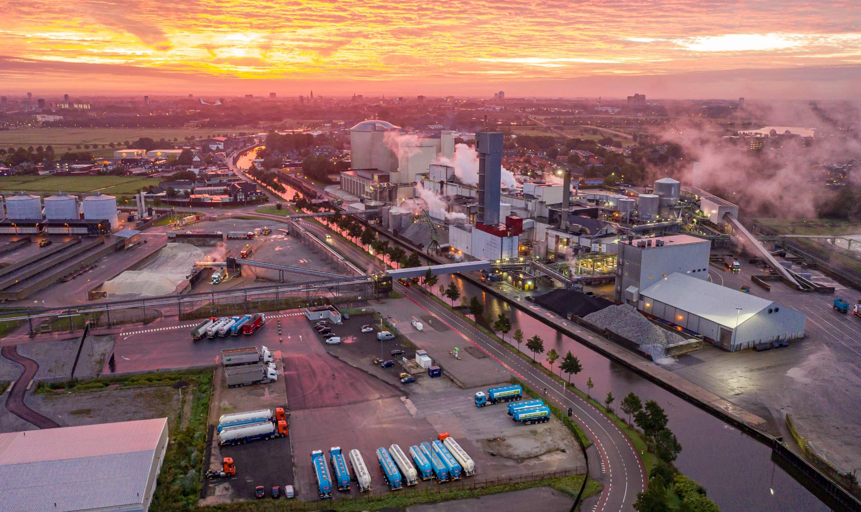 Aerial view of industrial site at sunrise with factories, trucks, and smoke near river and city.