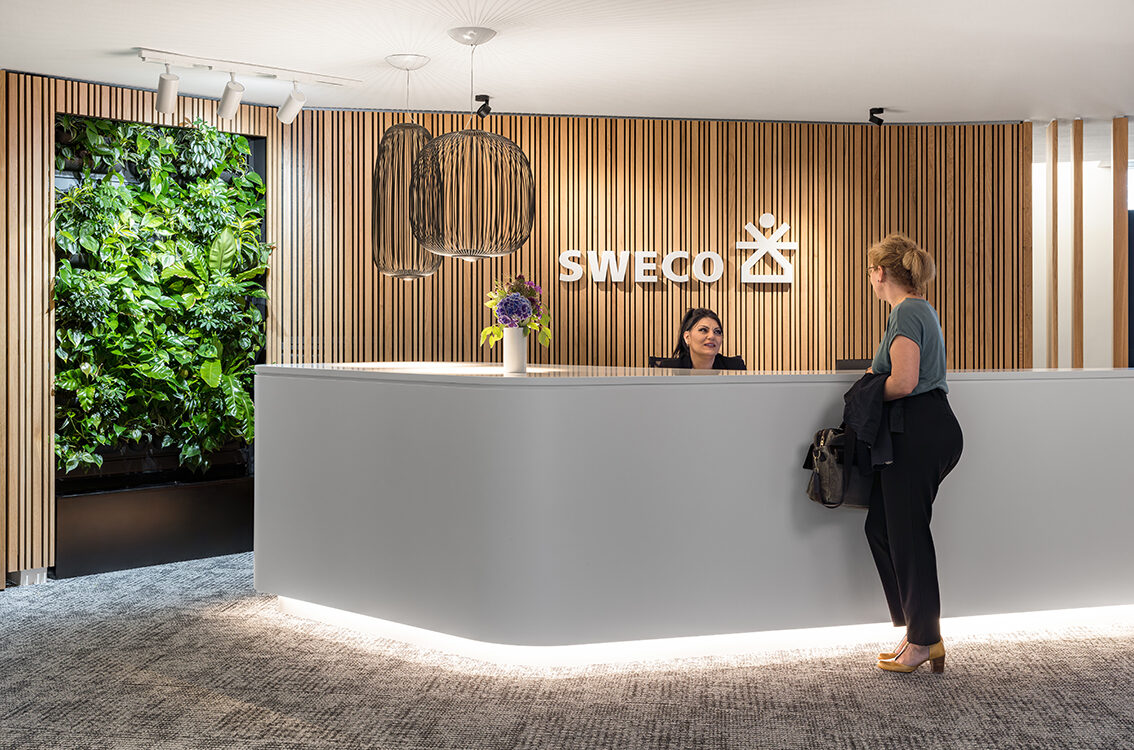 Modern office lobby, light wooden wall panels and white desk. Two middle-aged women discussing over the desk.