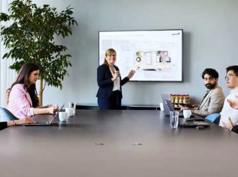 Iso neuvotteluhuoneen pöytä toimistossa, pöydän päässä keski-ikäinen nainen seisoo pitämässä esitystä ison näytön edessä. Pöydän vasemmalla puolella istuu kaksi naista, oikealla puolella kolme miestä.