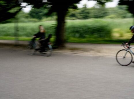 Kolme pyöräilijää vilahtaa oikealta vasemmalle, pyörätien varrella pyöräilijöiden takana näkyy lehtipuukuja ja niitty.