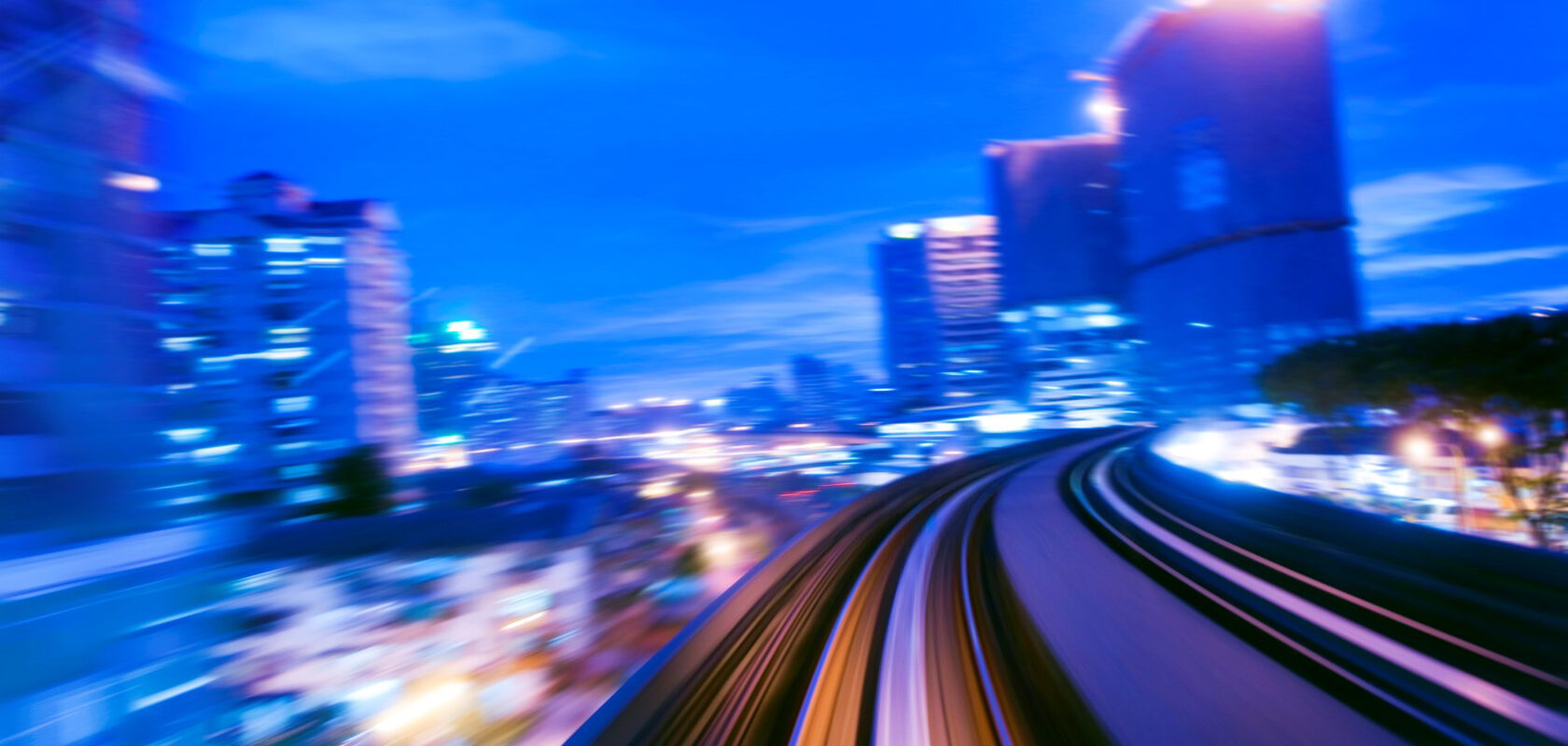 Nopeudella kuvattu kaupunkimaisema sinisen illan aikaan, jossa kaartu‑vat junaraiteet tai metrorata kulkevat korkeiden valaistujen rakennusten ohi ja valot muodostavat liike-epäterävää valojuovaa.
