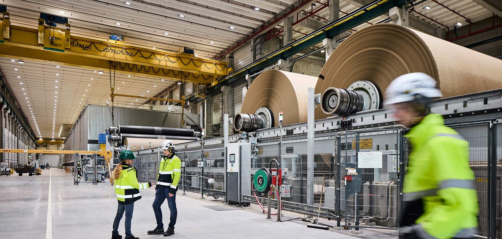 Workers in high visibility gear inspecting large paper rolls in modern factory.