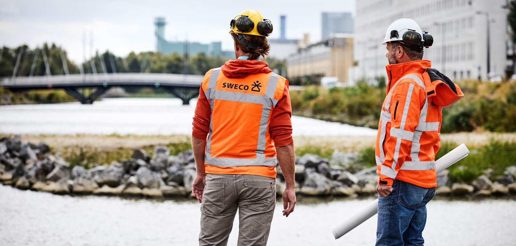 Engineers in orange safety gear inspecting riverside construction site near bridge.