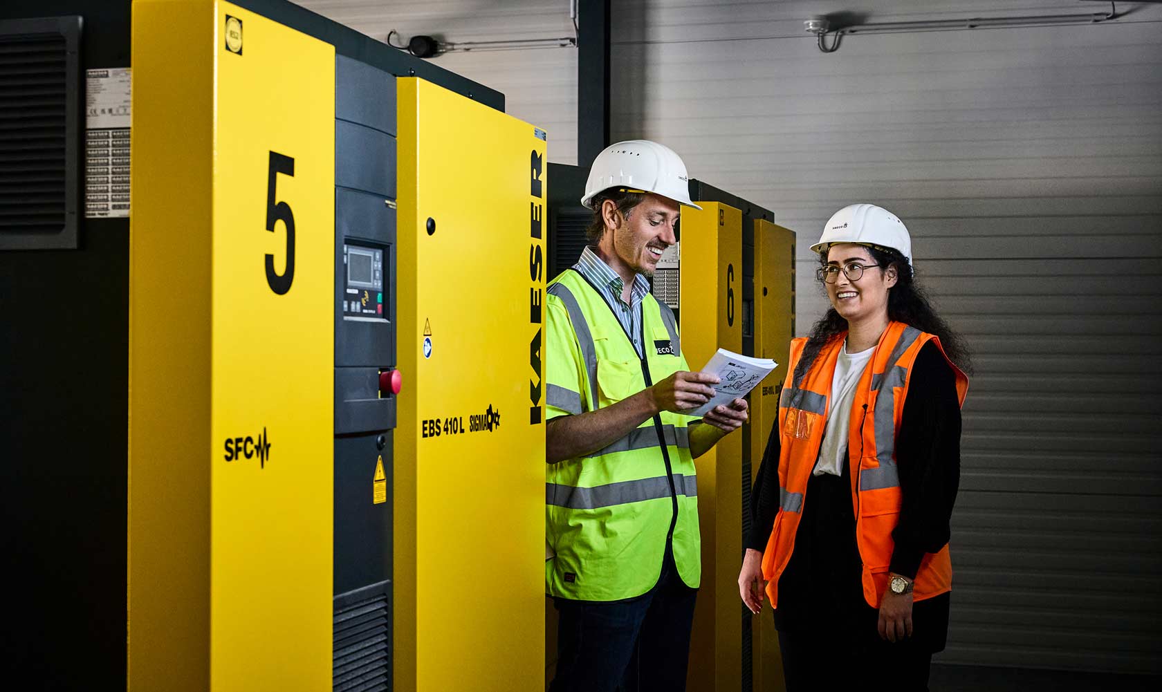 Engineers in safety vests inspect large yellow industrial compressors indoors.