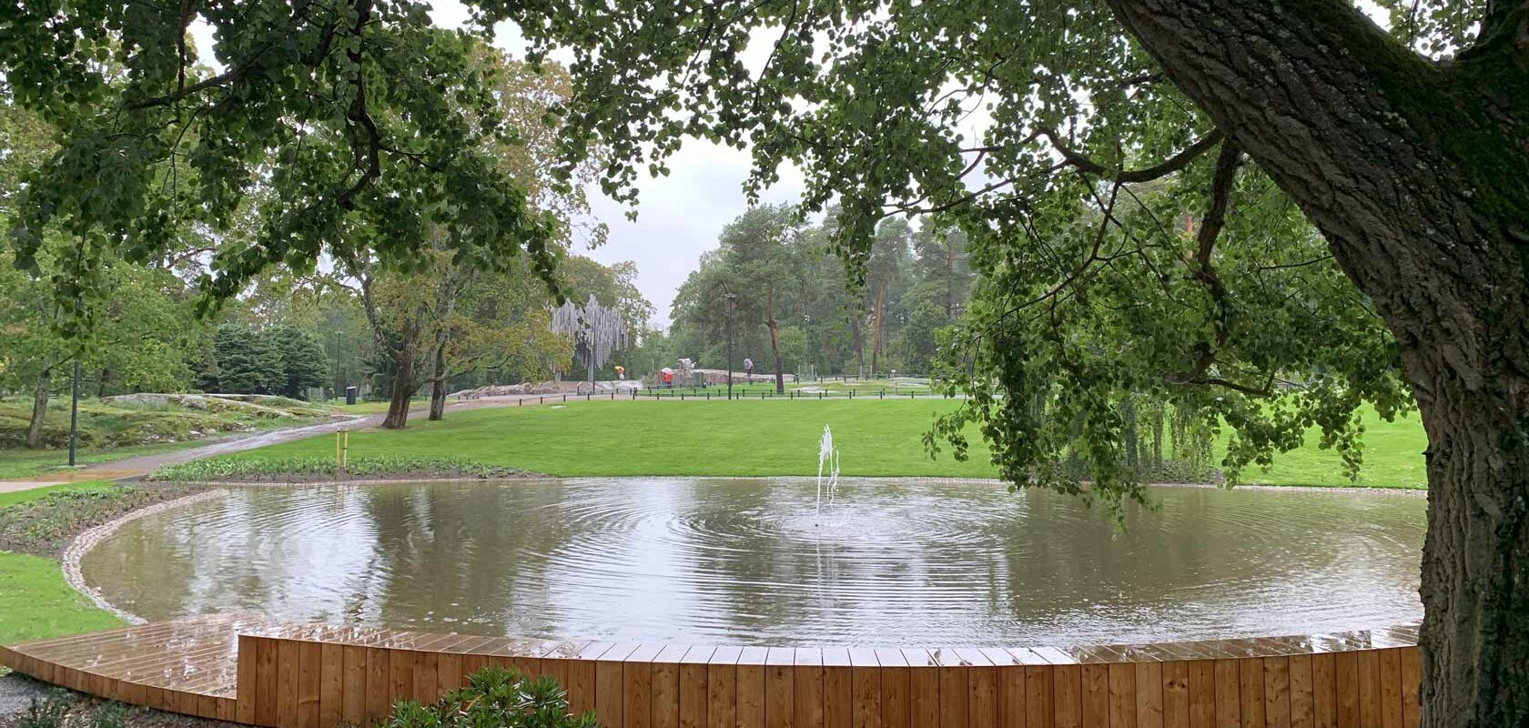 Tranquil park pond with wooden deck, fountain, and lush green trees in the background.