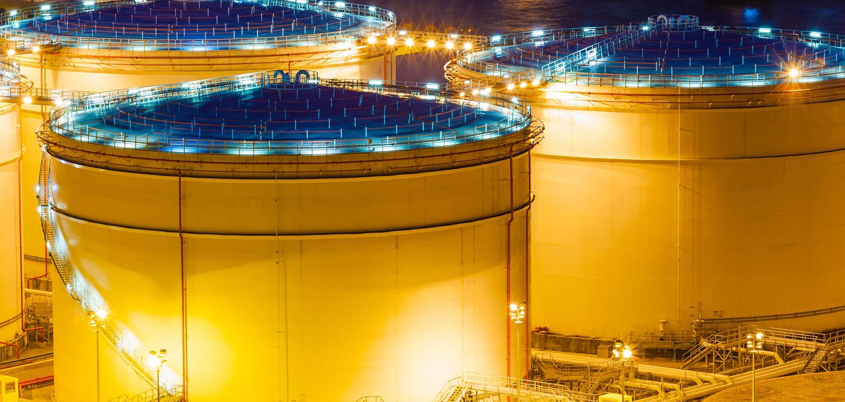 Large illuminated oil storage tanks at night in an industrial refinery complex.
