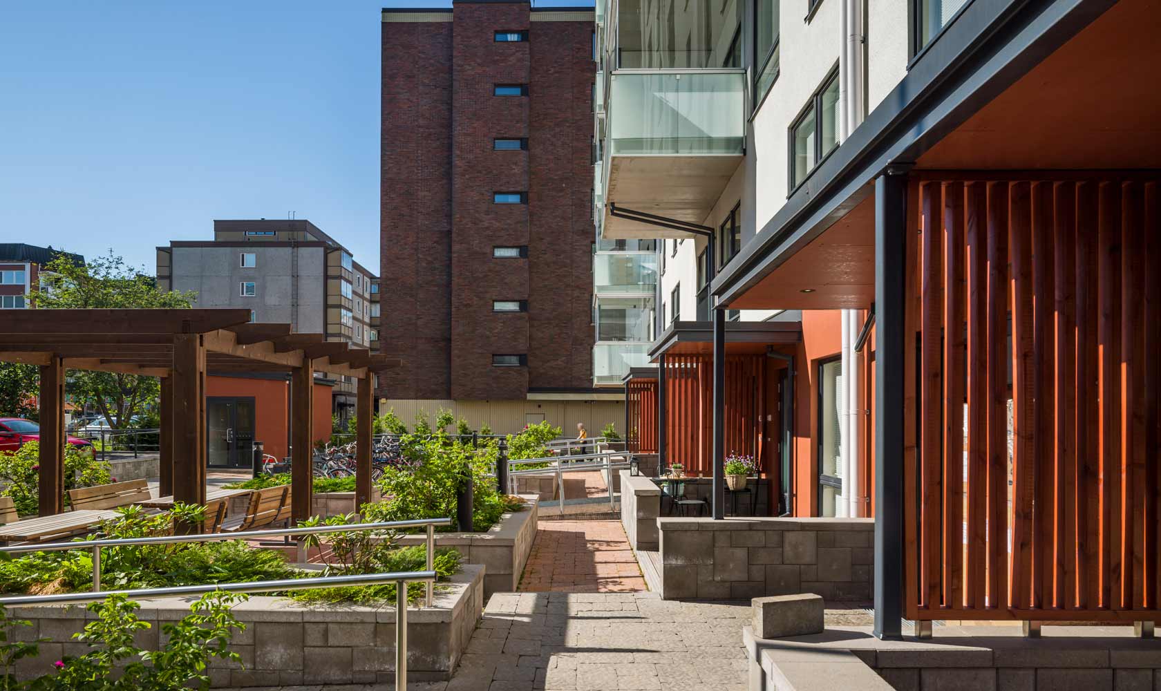 Moderni kerrostalon sisäpiha, pergola, istutusalueet ja polkupyörätelineet aurinkoisena päivänä.