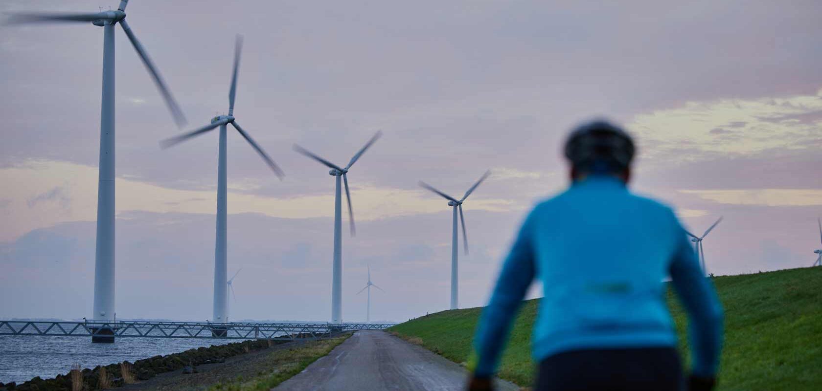 Cyclist riding on coastal road past modern wind turbines at sunset.