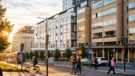 Modernia kaupunkirakennetta ilta-auringossa. Kadun varrella on kerrostaloja ja toimistorakennuksia, raitiotiekiskot, liikennevalot sekä jalankulkijoita ja pyöräilijöitä. Taustalla näkyy korkea tornitalo ja etualalla puu auringonvalossa.