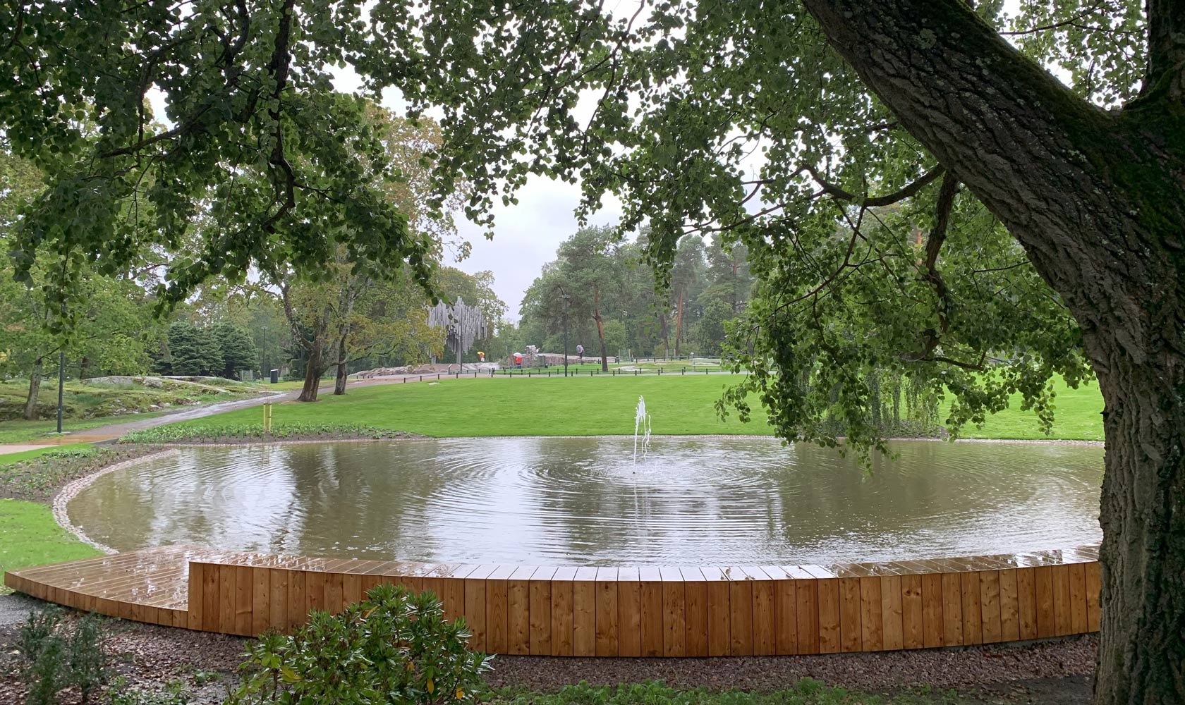Park pond with small fountain, wooden deck edge, and leafy trees on a rainy day.