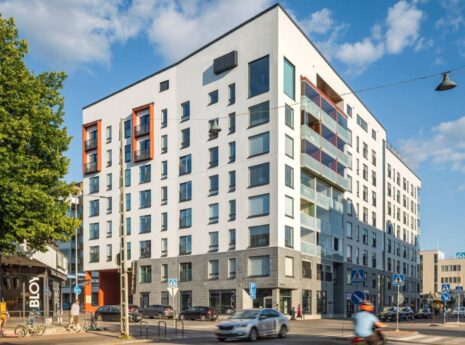 Moderni kerrostalokortteli kaupunkiympäristössä. Vaalea asuinrakennus sijaitsee kadunkulmassa, ja siinä on lasitettuja parvekkeita sekä suuria ikkunoita. Rakennuksen ympärillä on puita, liikennettä, pyöräilijöitä ja jalankulkijoita aurinkoisena päivänä.