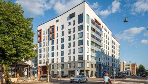 Moderni kerrostalokortteli kaupunkiympäristössä. Vaalea asuinrakennus sijaitsee kadunkulmassa, ja siinä on lasitettuja parvekkeita sekä suuria ikkunoita. Rakennuksen ympärillä on puita, liikennettä, pyöräilijöitä ja jalankulkijoita aurinkoisena päivänä.