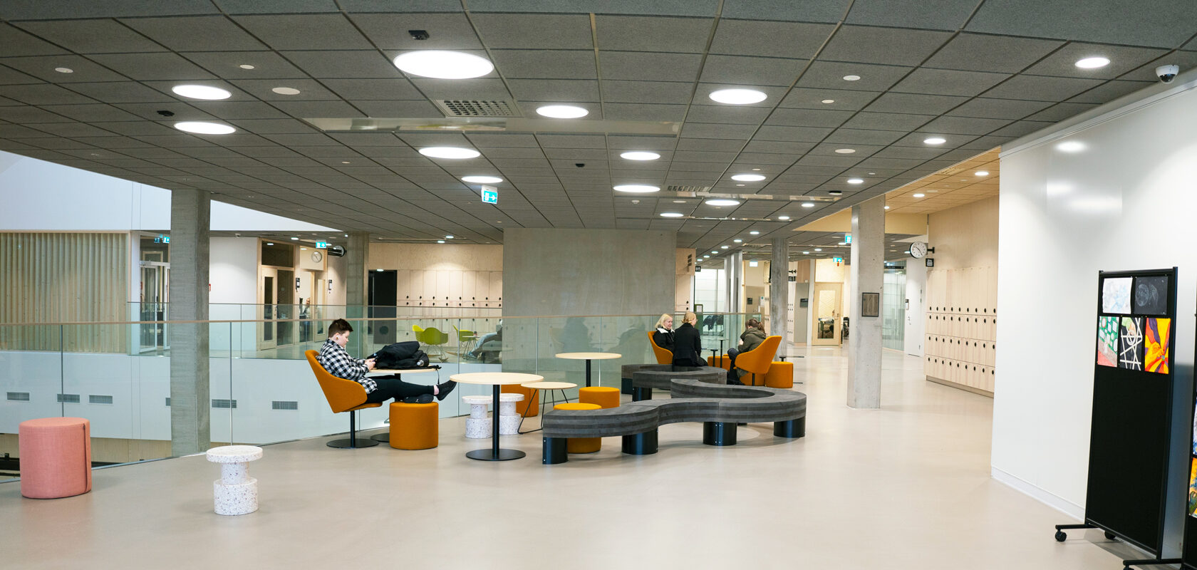 Modern university lounge with students on orange chairs, tables, and curved bench seating.