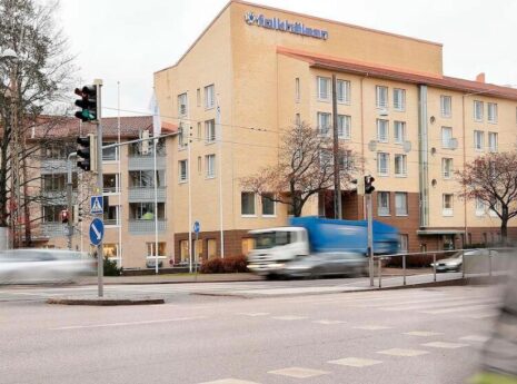 Keltainen Folkhälsan -talosta kuvattuna Mannerheimintieltä, etualalla liikkeestä blurraantunut jalankulkija.