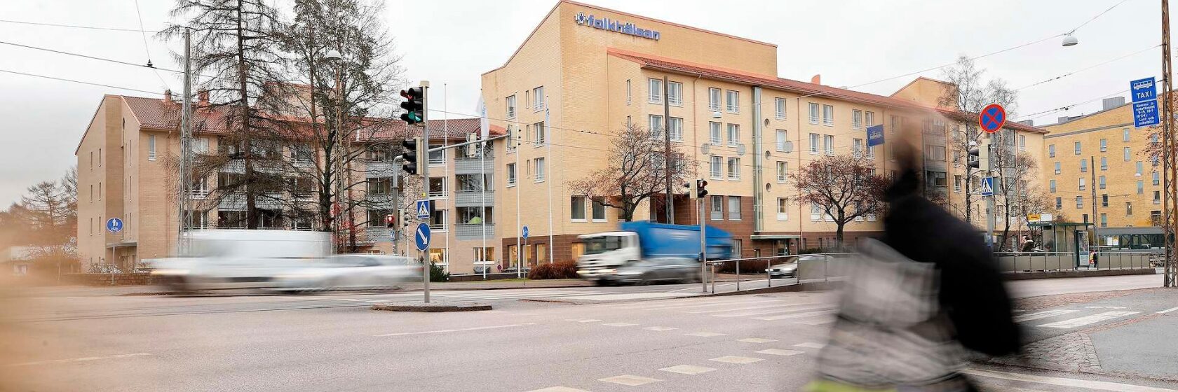 Keltainen Folkhälsan -talosta kuvattuna Mannerheimintieltä, etualalla liikkeestä blurraantunut jalankulkija.