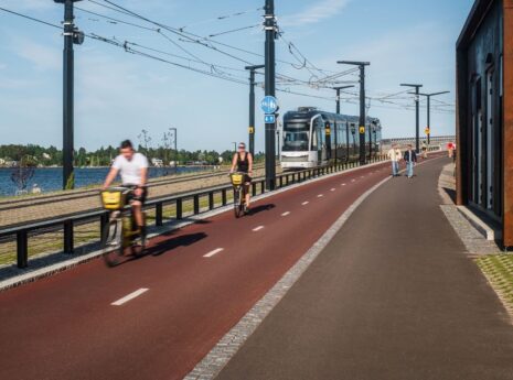 Raitiotie, pyörätie ja jalankulkuväylä kulkevat rinnakkain rantaympäristössä. Pyöräilijät, jalankulkijat ja raitiovaunu havainnollistavat eri kulkumuodoille suunniteltua liikenneympäristöä.