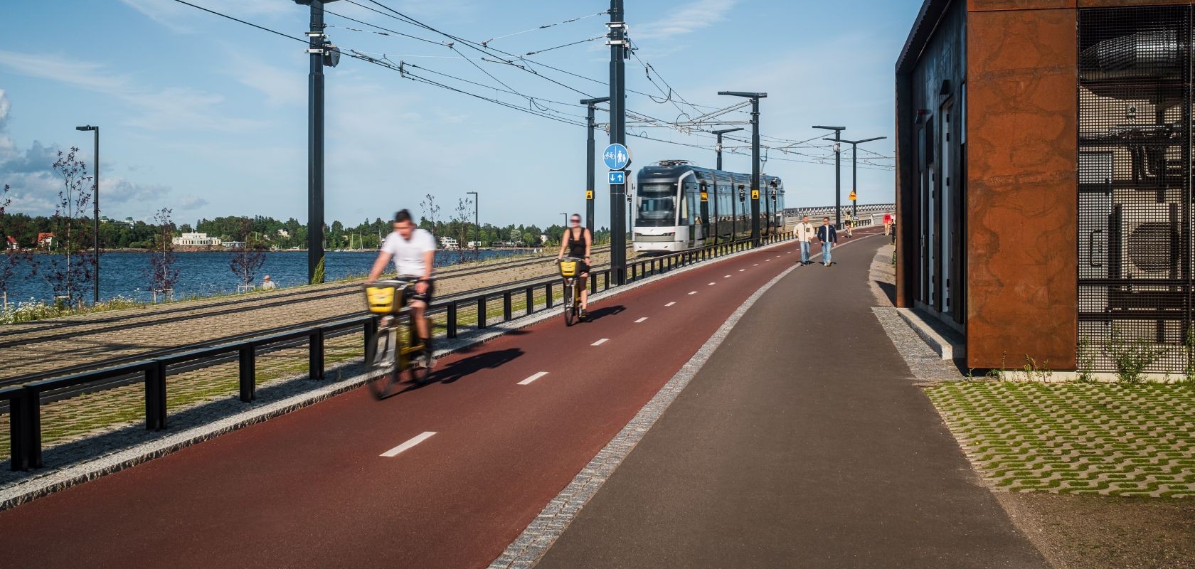 Raitiotie, pyörätie ja jalankulkuväylä kulkevat rinnakkain rantaympäristössä. Pyöräilijät, jalankulkijat ja raitiovaunu havainnollistavat eri kulkumuodoille suunniteltua liikenneympäristöä.