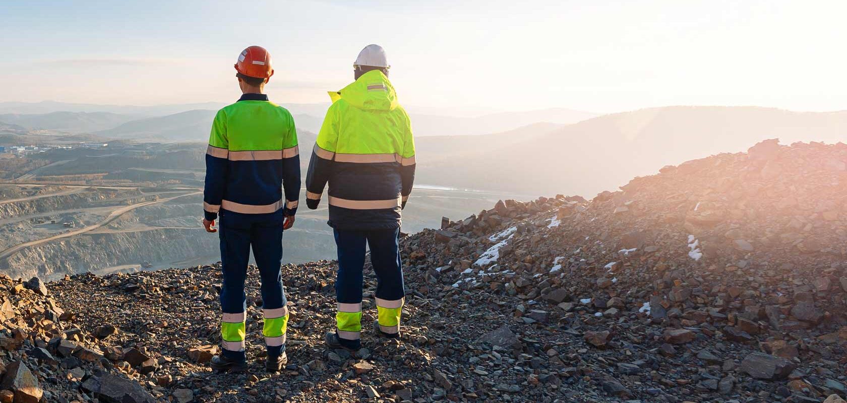 Two mining workers in safety gear overlooking an open pit mine at sunrise.