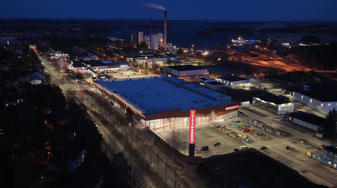 Pimeällä otettu ilmakuva valaistusta kauppakeskuksesta, taustalla teollisuusalue ja järvimaisema.