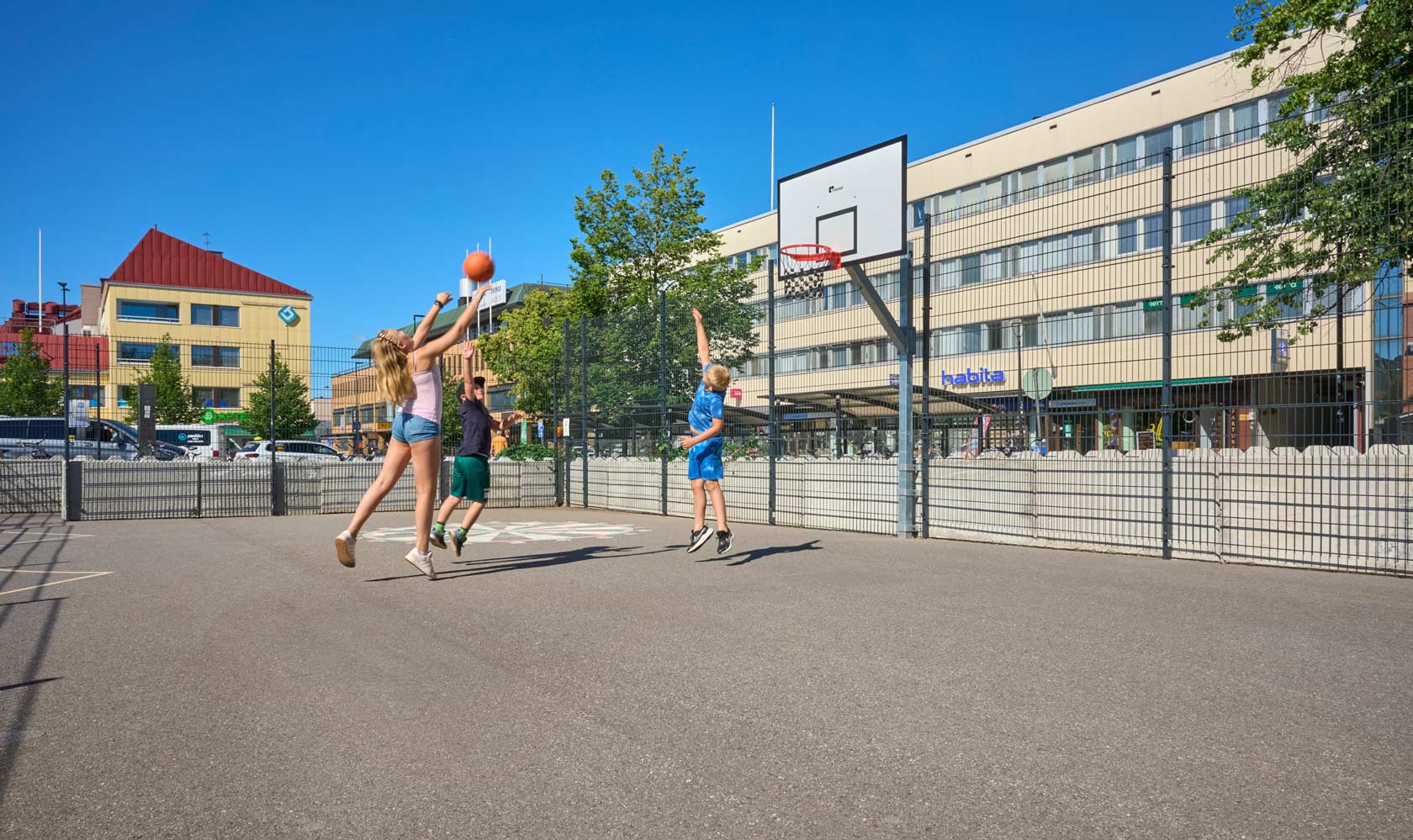 Kids playing basketball on an outdoor court with hoop and city buildings in background.