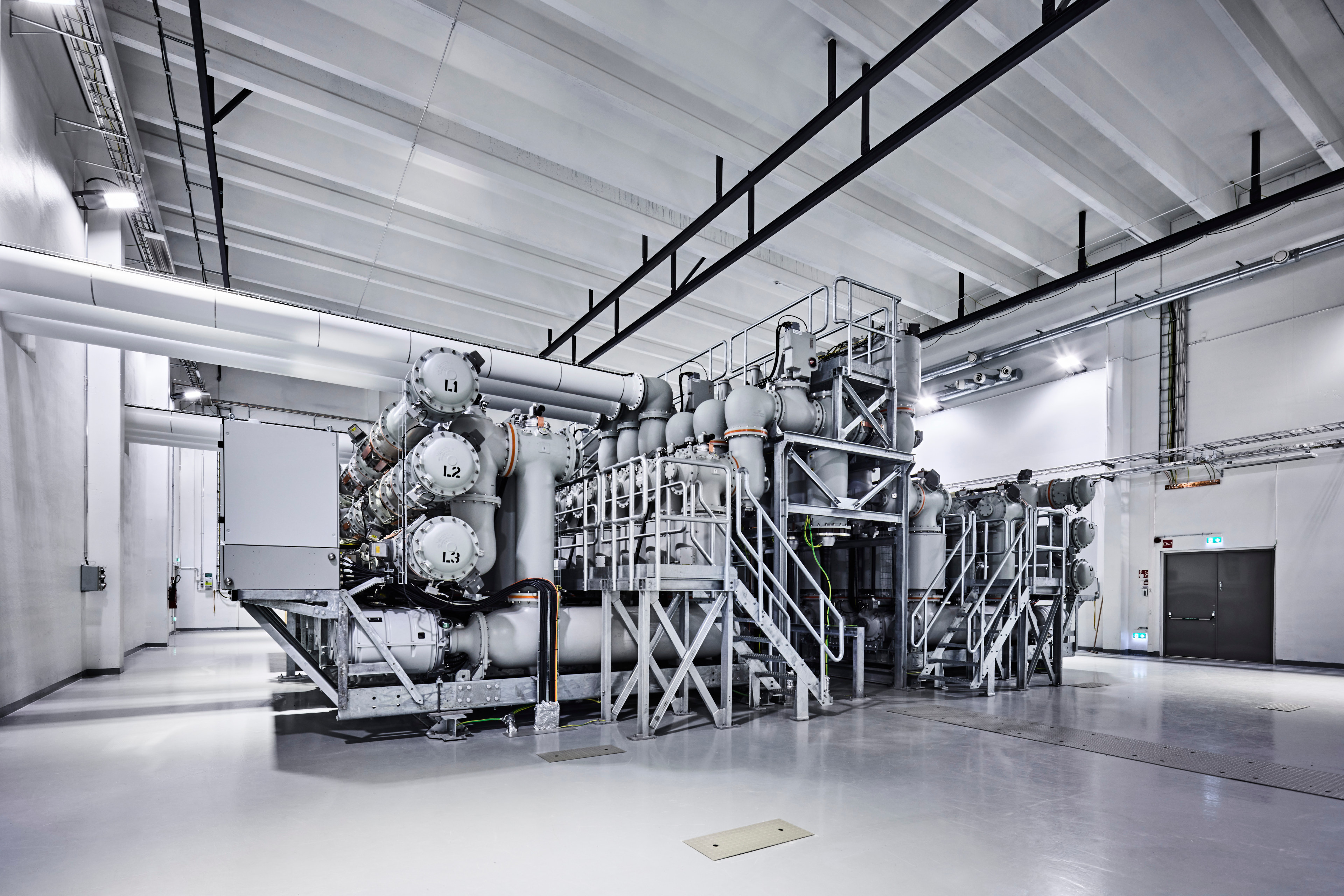 District heating network - substation hall. Tubular modules where the phases are marked L1–L3. There is metal maintenance platforms and stairs around the equipment.