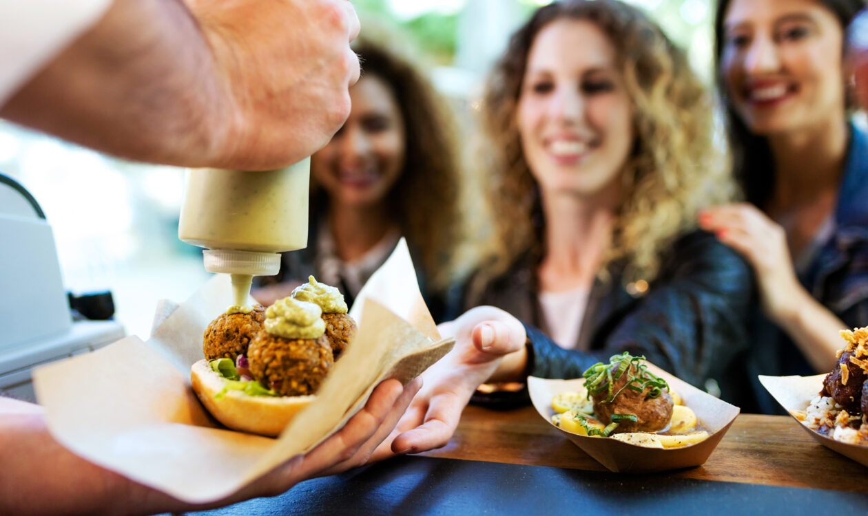 Street food vendor adding sauce to falafel sandwich as customers smile at counter.