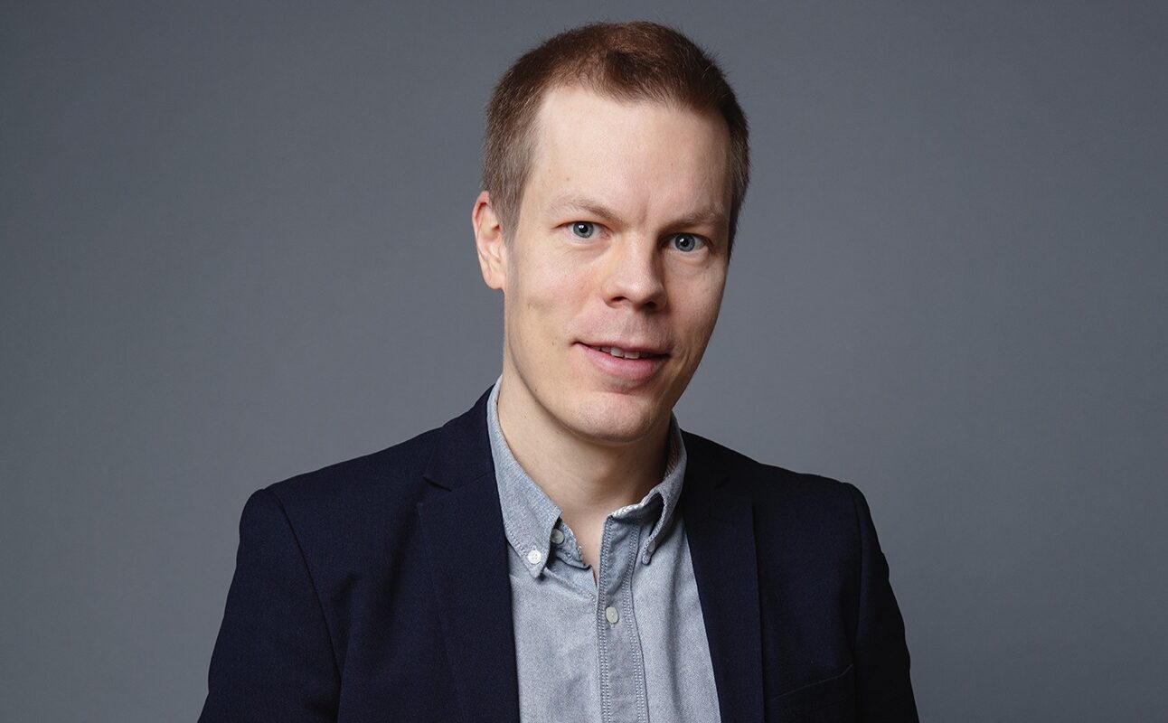 A studio portrait of a white male with short hair, wearing dark blue bleiser and gray shirt.