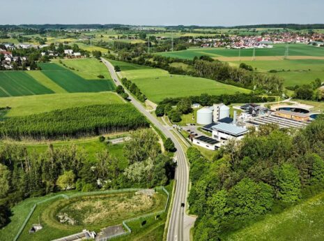 Ilmakuva vehreästä peltomaisemasta, taka-alalla siintää pieni matalarakenteinen kaupunki. Kuvaa halkoo maantie, joka johtaa kuvan edustalta kohti kaupunkia. Tien varressa on vesihuoltolaitos ja vedenpuhdistamo.